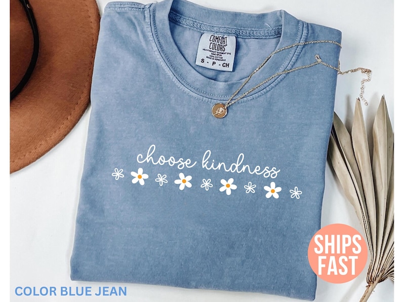 May include: A blue jean colored t-shirt with the words "choose kindness" in white script, accompanied by white and orange daisy graphics. A gold necklace with a pendant rests on the shirt.