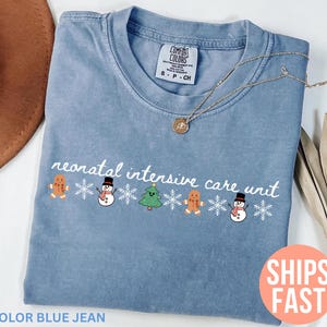 May include: A blue jean colored t-shirt with the text "neonatal intensive care unit" in white script. The shirt features a festive design of gingerbread men, snowmen, and a Christmas tree. A gold necklace rests on the shirt.