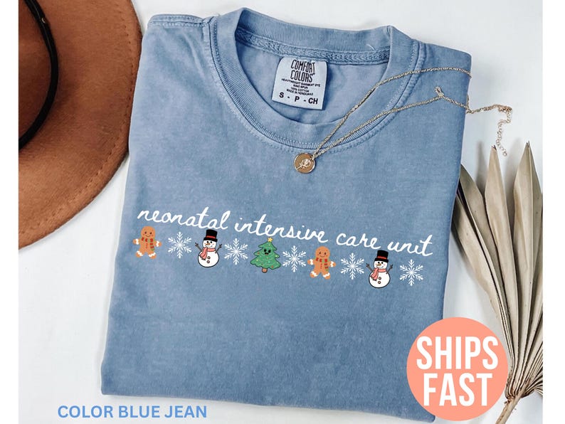 May include: A blue jean colored t-shirt with the text "neonatal intensive care unit" in white script. The shirt features a festive design of gingerbread men, snowmen, and a Christmas tree. A gold necklace rests on the shirt.