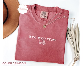 May include: A crimson short-sleeve t-shirt featuring the white text "wee woo crew" above a simple line-art graphic of an ambulance. The shirt has a crew neckline and a Comfort Colors brand label printed on the inside of the collar.