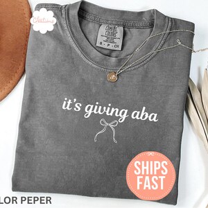 May include: A charcoal gray short-sleeve t-shirt with a crew neckline. The shirt features the white text "it's giving aba" printed on the chest, accompanied by a small white line-art bow graphic positioned directly below the text.