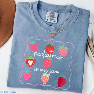 May include: A blue jean coloured t-shirt with the phrase "pediatrics is my jam" in white script, surrounded by a pink frame and strawberry illustrations. A gold necklace with a pendant is draped across the shirt.