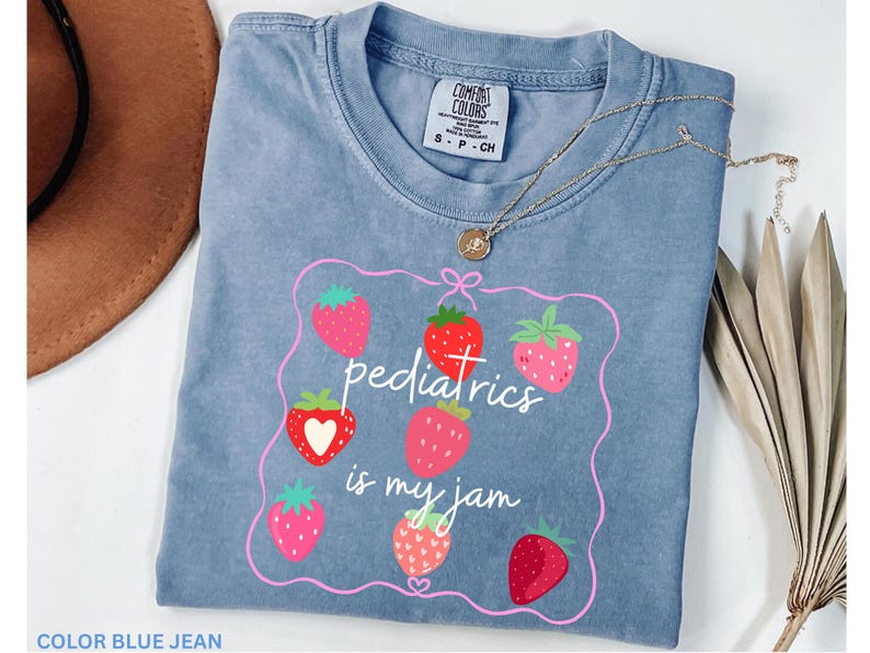 May include: A blue jean coloured t-shirt with the phrase "pediatrics is my jam" in white script, surrounded by a pink frame and strawberry illustrations. A gold necklace with a pendant is draped across the shirt.