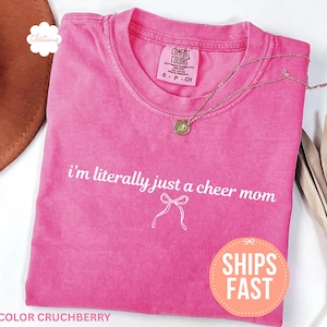 May include: A pink t-shirt with the text "i'm literally just a cheer mom" in white. A gold necklace and a brown hat are also visible. The shirt has a round neck and short sleeves. The shirt is labeled "Color Cruchberry".