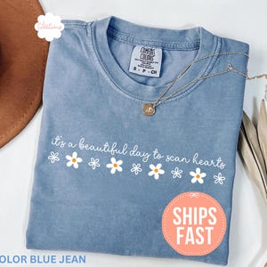 以下が含まれることがあります： 「it's a beautiful day to scan hearts」の文字とデイジーのグラフィックが入った、ブルーデニムカラーのTシャツ。ゴールドのネックレスと茶色の帽子が見えます。シャツにはComfort Colorsのタグが付いています。