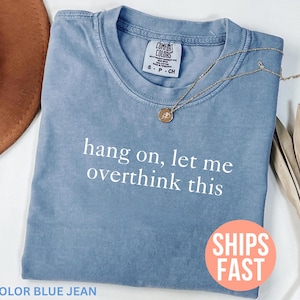 Pode incluir: Camiseta azul jeans com a frase branca "hang on, let me overthink this". Um colar de ouro com um pingente repousa sobre a camisa. Um chapéu marrom e uma planta seca estão no fundo.
