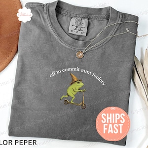 May include: A gray Comfort Colors t-shirt with the phrase "off to commit aunt foolery" and a graphic of a frog riding a scooter. The frog wears a hat and glasses. A necklace and a "Ships Fast" sticker are also visible.