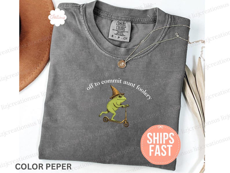May include: A gray Comfort Colors t-shirt with the phrase "off to commit aunt foolery" and a graphic of a frog riding a scooter. The frog wears a hat and glasses. A necklace and a "Ships Fast" sticker are also visible.