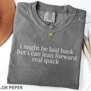 May include: A gray Comfort Colors t-shirt with the text "i might be laid back but i can lean forward real quick" in white. A gold pendant necklace and a brown hat are also visible. The shirt is folded.
