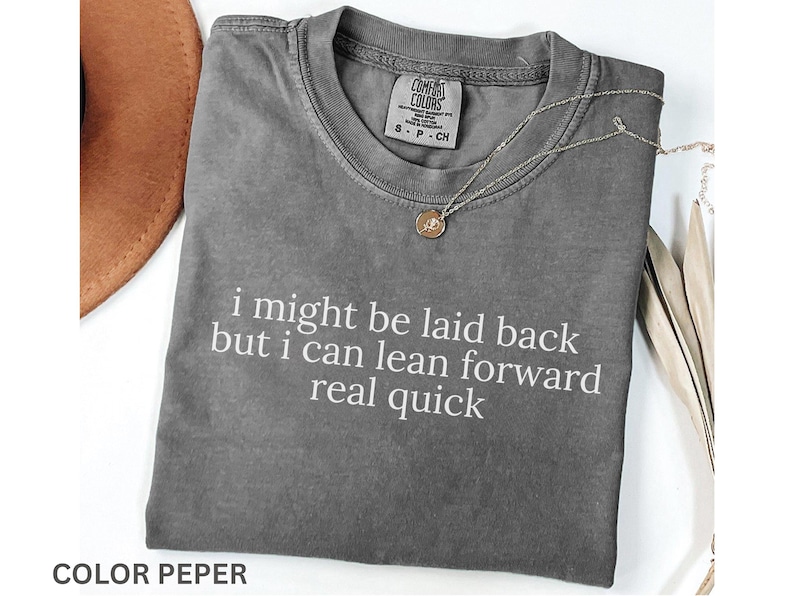 May include: A gray Comfort Colors t-shirt with the text "i might be laid back but i can lean forward real quick" in white. A gold pendant necklace and a brown hat are also visible. The shirt is folded.