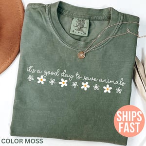 K&ouml;nnte beinhalten: Olivgr&uuml;nes T-Shirt mit dem wei&szlig;en Schriftzug "it's a good day to save animals" und G&auml;nsebl&uuml;mchen-Grafiken. Das Shirt ist von der Marke Comfort Colors und die Farbe ist "Moss". Eine goldene Halskette liegt auf dem Shirt.