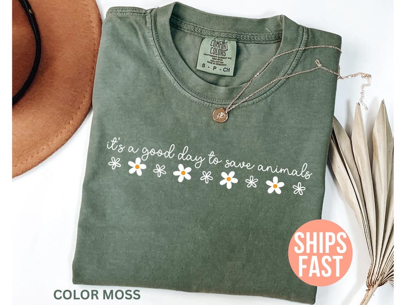 K&ouml;nnte beinhalten: Olivgr&uuml;nes T-Shirt mit dem wei&szlig;en Schriftzug "it's a good day to save animals" und G&auml;nsebl&uuml;mchen-Grafiken. Das Shirt ist von der Marke Comfort Colors und die Farbe ist "Moss". Eine goldene Halskette liegt auf dem Shirt.