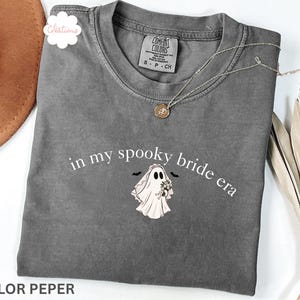 May include: A pepper gray t-shirt featuring the white text "in my spooky bride era" arched above a graphic of a ghost wearing a bridal veil and holding a bouquet of flowers, with two small black bats flanking the ghost's head.