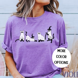 May include: Violet-colored long-sleeve shirt featuring a playful Halloween design. The graphic depicts four cartoon cats dressed as ghosts, with one wearing a witch hat. The shirt has the words "MORE COLOR OPTIONS" and "VIOLET" on it.