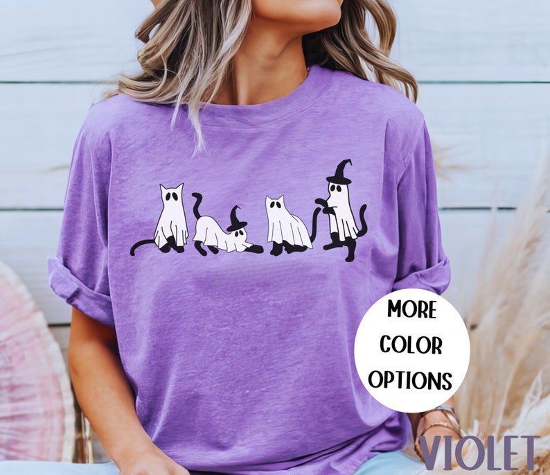 May include: Violet-colored long-sleeve shirt featuring a playful Halloween design. The graphic depicts four cartoon cats dressed as ghosts, with one wearing a witch hat. The shirt has the words "MORE COLOR OPTIONS" and "VIOLET" on it.
