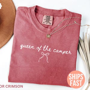 May include: Crimson-colored t-shirt with the text "queen of the camper" in white script. A gold necklace with a pendant rests on the shirt. The shirt is folded and placed on a white surface.