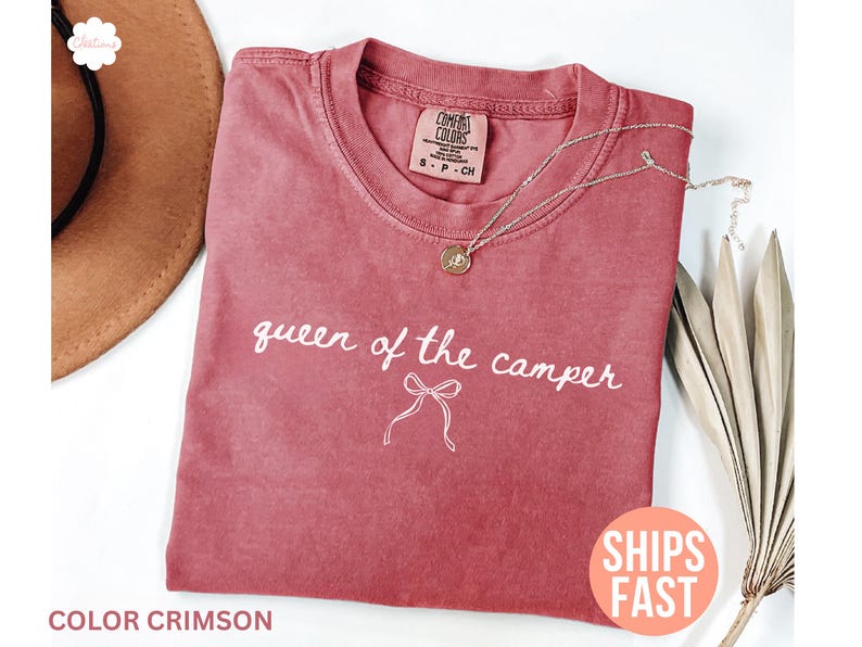 May include: Crimson-colored t-shirt with the text "queen of the camper" in white script. A gold necklace with a pendant rests on the shirt. The shirt is folded and placed on a white surface.