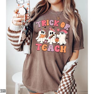 May include: Espresso-colored t-shirt with the words "Trick or Teach" in a retro font. The graphic features three cartoon ghosts, one reading a book, one holding a pencil, and one with a pumpkin bucket. The shirt has a relaxed fit.