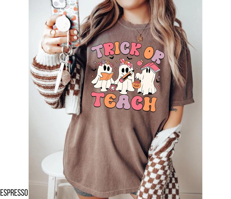 May include: Espresso-colored t-shirt with the words "Trick or Teach" in a retro font. The graphic features three cartoon ghosts, one reading a book, one holding a pencil, and one with a pumpkin bucket. The shirt has a relaxed fit.