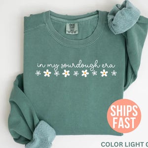 May include: Light green sweatshirt with the phrase "in my sourdough era" in white script, decorated with small daisy graphics. The sweatshirt is folded, with the sleeves and collar visible. The text "SHIPS FAST" is on a round orange sticker.