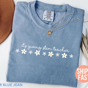 Puede incluir: Una camiseta azul vaquera con la frase "it's giving stem teacher" en escritura blanca, adornada con gráficos de margaritas. Un collar dorado con un colgante descansa sobre la camisa. La camiseta tiene una etiqueta Comfort Colors.