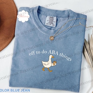 May include: A blue jean colored t-shirt featuring a graphic of a white goose holding a coffee cup. The text above the goose reads "off to do ABA things". The shirt has a crew neckline and a Comfort Colors brand label visible at the inner collar.