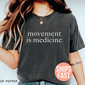 K&ouml;nnte beinhalten: Dunkelgraues T-Shirt mit dem wei&szlig;en Text "movement is medicine". Das Shirt hat eine einfarbige Farbe und einen Rundhalsausschnitt. Das Bild enth&auml;lt auch einen runden Aufkleber mit der Aufschrift "SHIPS FAST".