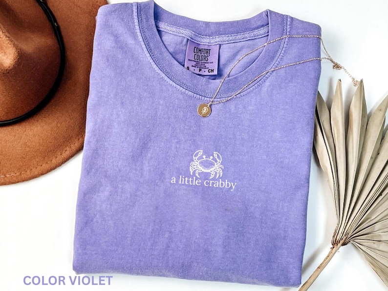 May include: A violet Comfort Colors t-shirt with a white crab graphic and the phrase "a little crabby." A gold necklace and a brown hat are also visible. The shirt is a casual style.
