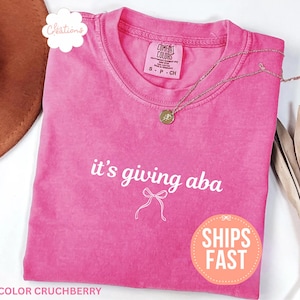 May include: A bright pink short-sleeve t-shirt with a crew neckline. The shirt features the white text 'it's giving aba' printed above a simple white line drawing of a bow. The garment has a Comfort Colors brand label at the inner neck.