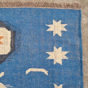Puede incluir: Alfombra tejida azul con un dise&ntilde;o geom&eacute;trico. La alfombra presenta un dise&ntilde;o central con una estrella naranja en un cuadrado crema, rodeado por un borde gris oscuro. La alfombra tambi&eacute;n tiene formas de estrella color crema y flecos.