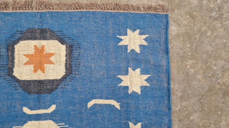 Puede incluir: Alfombra tejida azul con un dise&ntilde;o geom&eacute;trico. La alfombra presenta un dise&ntilde;o central con una estrella naranja en un cuadrado crema, rodeado por un borde gris oscuro. La alfombra tambi&eacute;n tiene formas de estrella color crema y flecos.