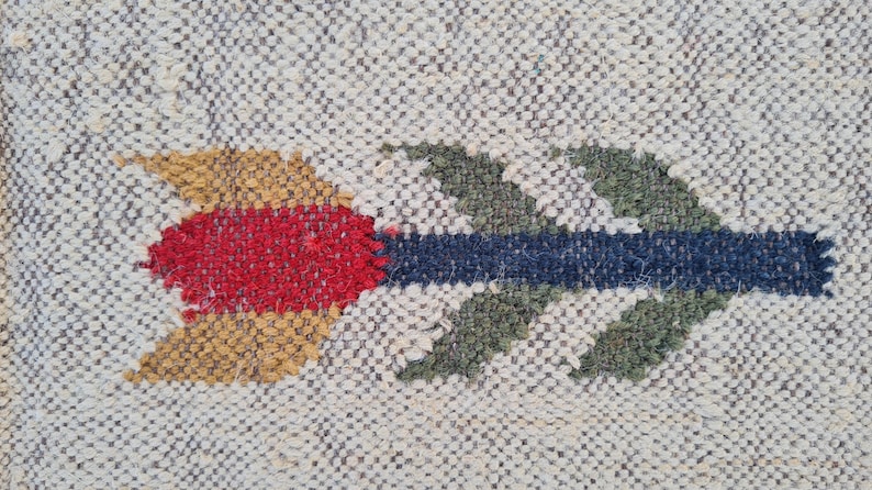 May include: Close-up of a woven textile with a repeating pattern. The design features a colorful arrow in shades of red, gold, blue, and green. The arrow is centered on a neutral background.