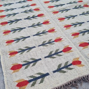 May include: A rectangular beige rug with a repeating floral pattern. The flowers are red and yellow with green stems. The rug has a fringed edge and is made of a woven material.