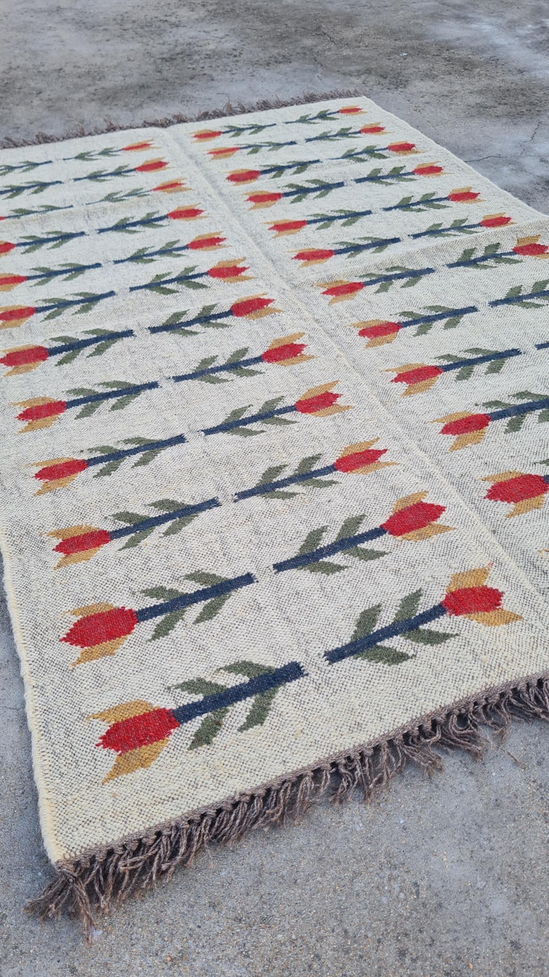 May include: A rectangular beige rug with a repeating floral pattern. The flowers are red and yellow with green stems. The rug has a fringed edge and is made of a woven material.