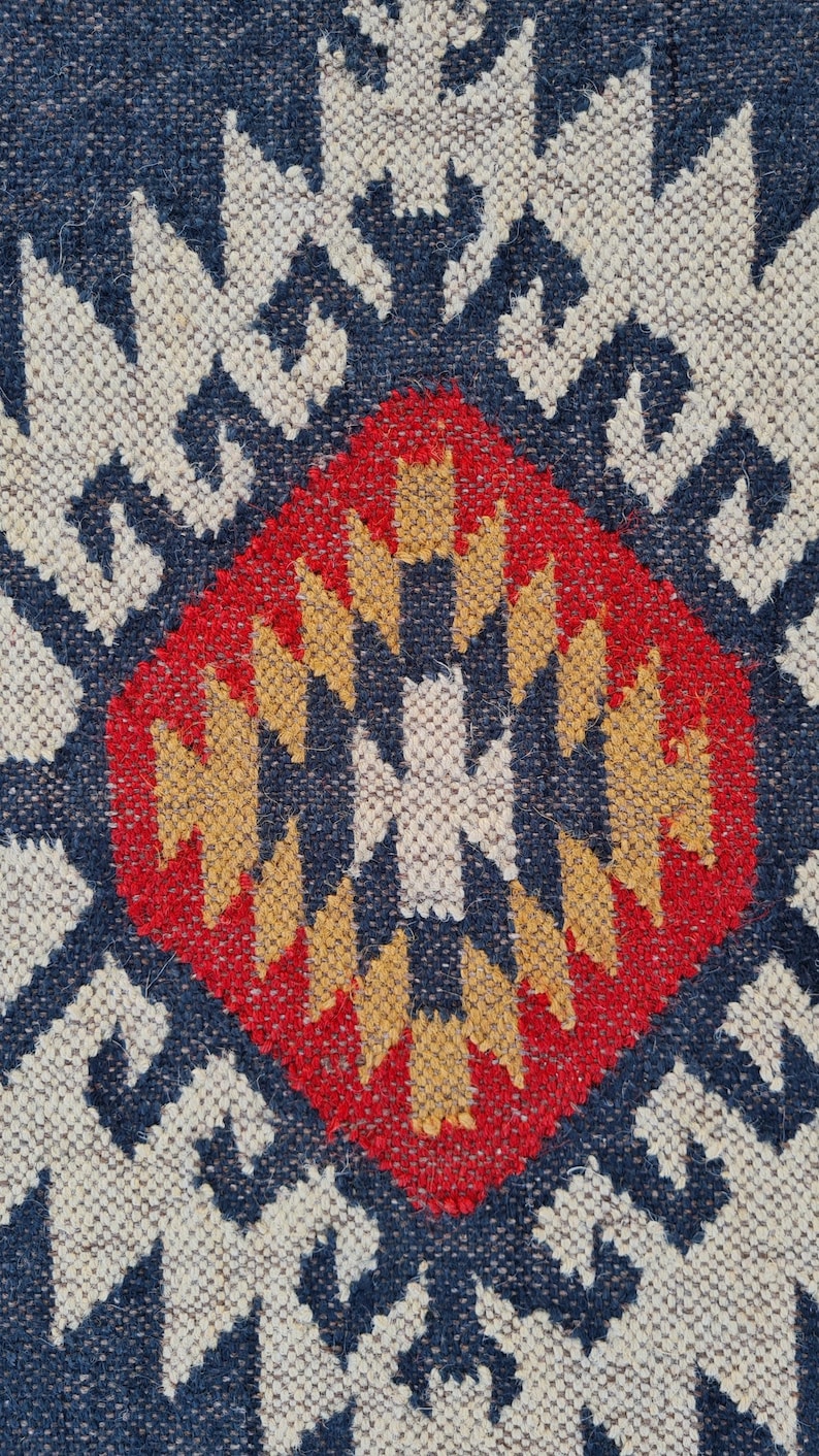 May include: Close-up of a woven rug with a geometric pattern. The rug features a central red diamond shape with yellow and white accents, surrounded by a navy blue and white design. The rug has a textured appearance.
