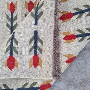 May include: Beige woven rug with a repeating floral pattern. The design features red, yellow, and green stylized flowers with blue stems. The rug has a fringed edge and is partially rolled up, revealing the underside.