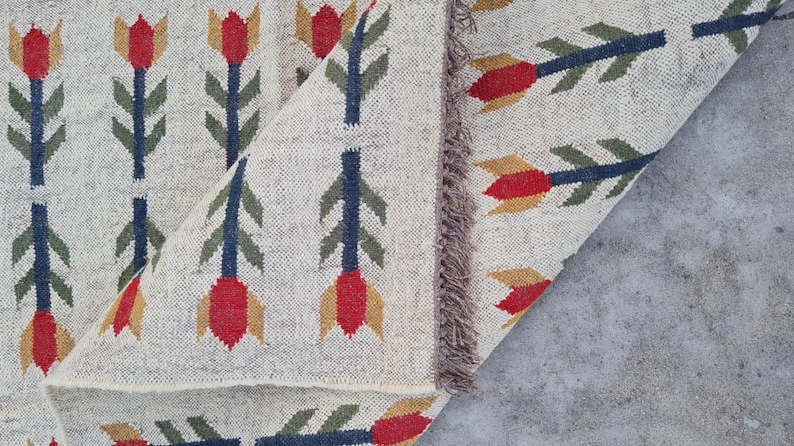 May include: Beige woven rug with a repeating floral pattern. The design features red, yellow, and green stylized flowers with blue stems. The rug has a fringed edge and is partially rolled up, revealing the underside.
