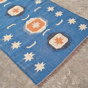Puede incluir: Una alfombra rectangular azul con un patr&oacute;n repetido de estrellas blancas, formas geom&eacute;tricas naranjas y blancas y l&iacute;neas curvas blancas. La alfombra tiene un borde con flecos y est&aacute; hecha de un material tejido.