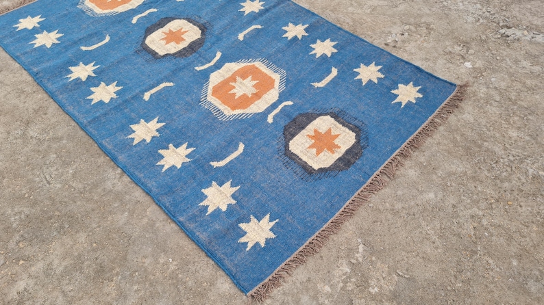 Puede incluir: Una alfombra rectangular azul con un patr&oacute;n repetido de estrellas blancas, formas geom&eacute;tricas naranjas y blancas y l&iacute;neas curvas blancas. La alfombra tiene un borde con flecos y est&aacute; hecha de un material tejido.