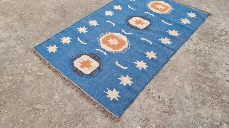 Puede incluir: Una alfombra rectangular azul con un patr&oacute;n repetido de estrellas blancas, formas de media luna y dise&ntilde;os florales estilizados en naranja y blanco. La alfombra tiene un fleco marr&oacute;n en los bordes.