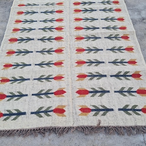May include: Cream-colored woven rug with a repeating pattern of red, green, and yellow floral arrow designs. The rug has a fringed edge and is displayed on a concrete surface. A decorative floor covering.