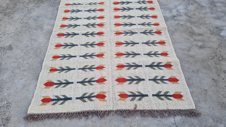 May include: Cream-colored woven rug with a repeating pattern of red, green, and yellow floral arrow designs. The rug has a fringed edge and is displayed on a concrete surface. A decorative floor covering.