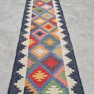 Puede incluir: Una alfombra de pasillo rectangular y colorida con un patr&oacute;n geom&eacute;trico. La alfombra presenta un borde azul oscuro, un borde en zigzag blanco y un dise&ntilde;o central de diamantes en varios colores, incluyendo rojo, amarillo, azul y verde.