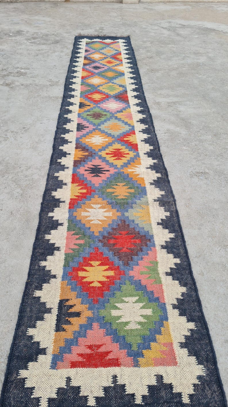 Puede incluir: Una alfombra de pasillo rectangular y colorida con un patr&oacute;n geom&eacute;trico. La alfombra presenta un borde azul oscuro, un borde en zigzag blanco y un dise&ntilde;o central de diamantes en varios colores, incluyendo rojo, amarillo, azul y verde.