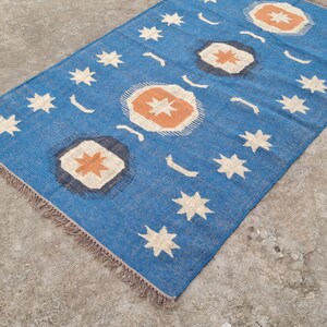 Puede incluir: Una alfombra rectangular azul con un patr&oacute;n repetido de estrellas blancas y formas florales estilizadas en naranja y blanco. La alfombra tiene un fleco marr&oacute;n. Esta alfombra decorativa es adecuada para una sala de estar o un dormitorio.