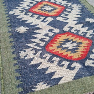 May include: A rectangular rug with a geometric pattern. The rug features a navy blue background with white and red diamond shapes. The border is green, and the rug has fringe on the ends. The rug is made of woven material.