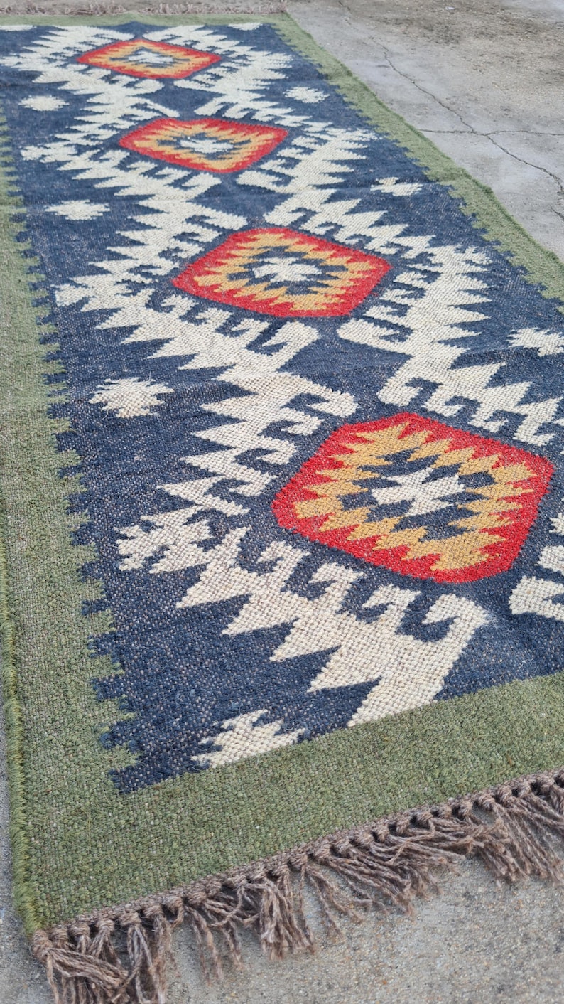 May include: A rectangular rug with a geometric pattern. The rug features a navy blue background with white and red diamond shapes. The border is green, and the rug has fringe on the ends. The rug is made of woven material.