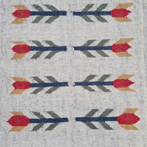 May include: A rectangular, off-white rug with a repeating floral pattern. The flowers are red and yellow with green leaves and blue stems. The rug has a fringed edge.