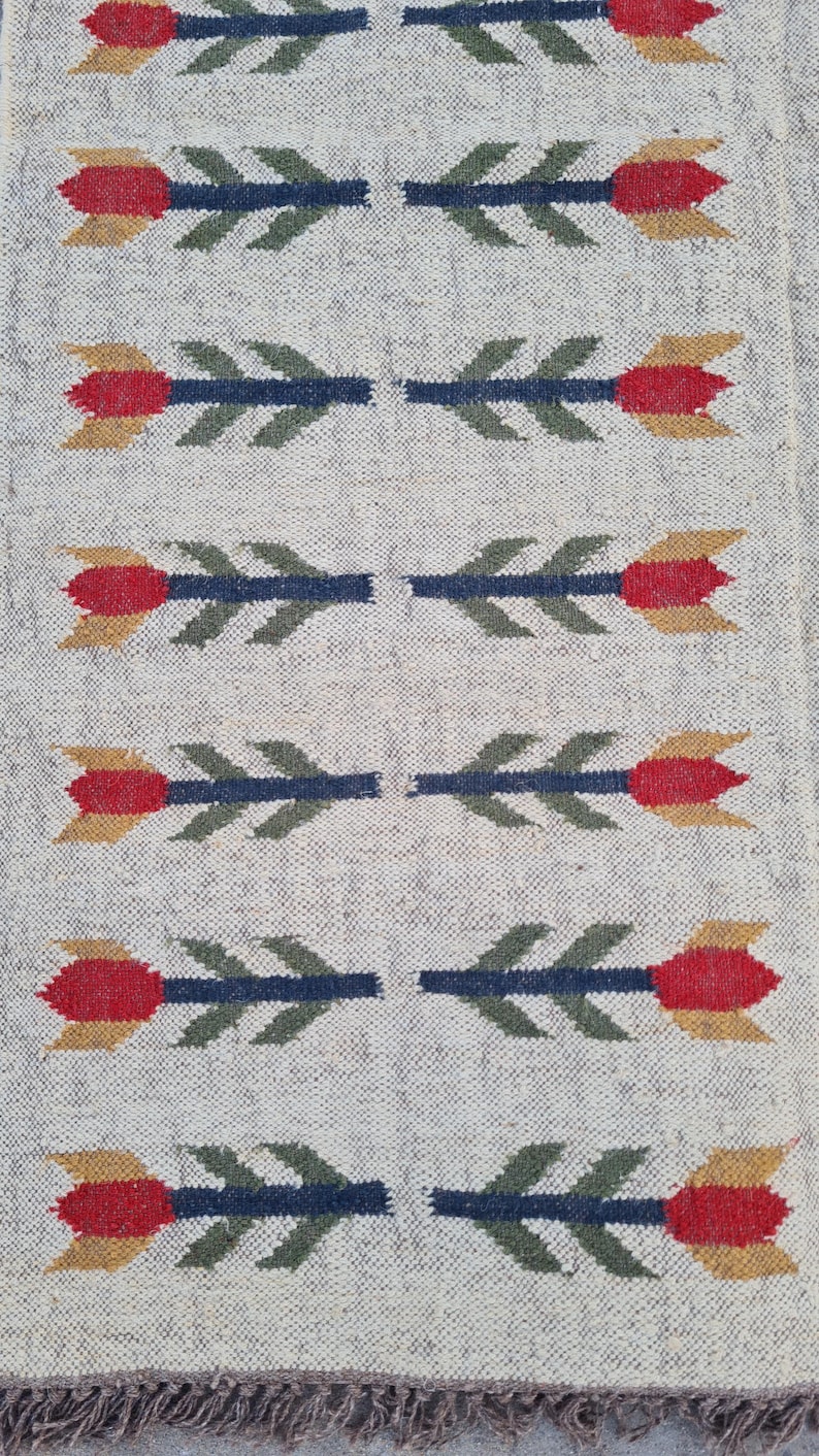 May include: A rectangular, off-white rug with a repeating floral pattern. The flowers are red and yellow with green leaves and blue stems. The rug has a fringed edge.