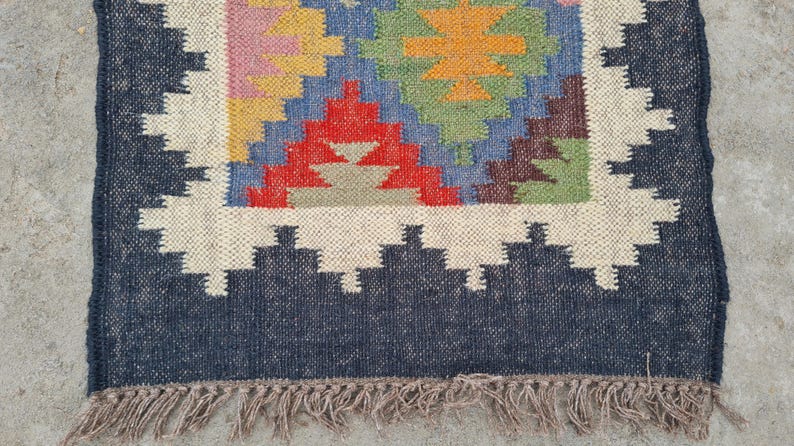 Puede incluir: Alfombra de yute tejida a mano con un patr&oacute;n geom&eacute;trico en rojo, azul, amarillo y verde. La alfombra tiene un borde azul oscuro con un dise&ntilde;o en zigzag blanco y un borde con flecos. Un revestimiento de suelo decorativo.